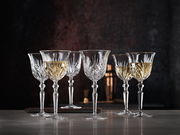 Sechs elegante Weißweinkelche aus Kristallglas mit feinem Schliffmuster, gefüllt mit Weißwein, auf einer Tischoberfläche vor dunklem Hintergrund, seitliche Perspektive.