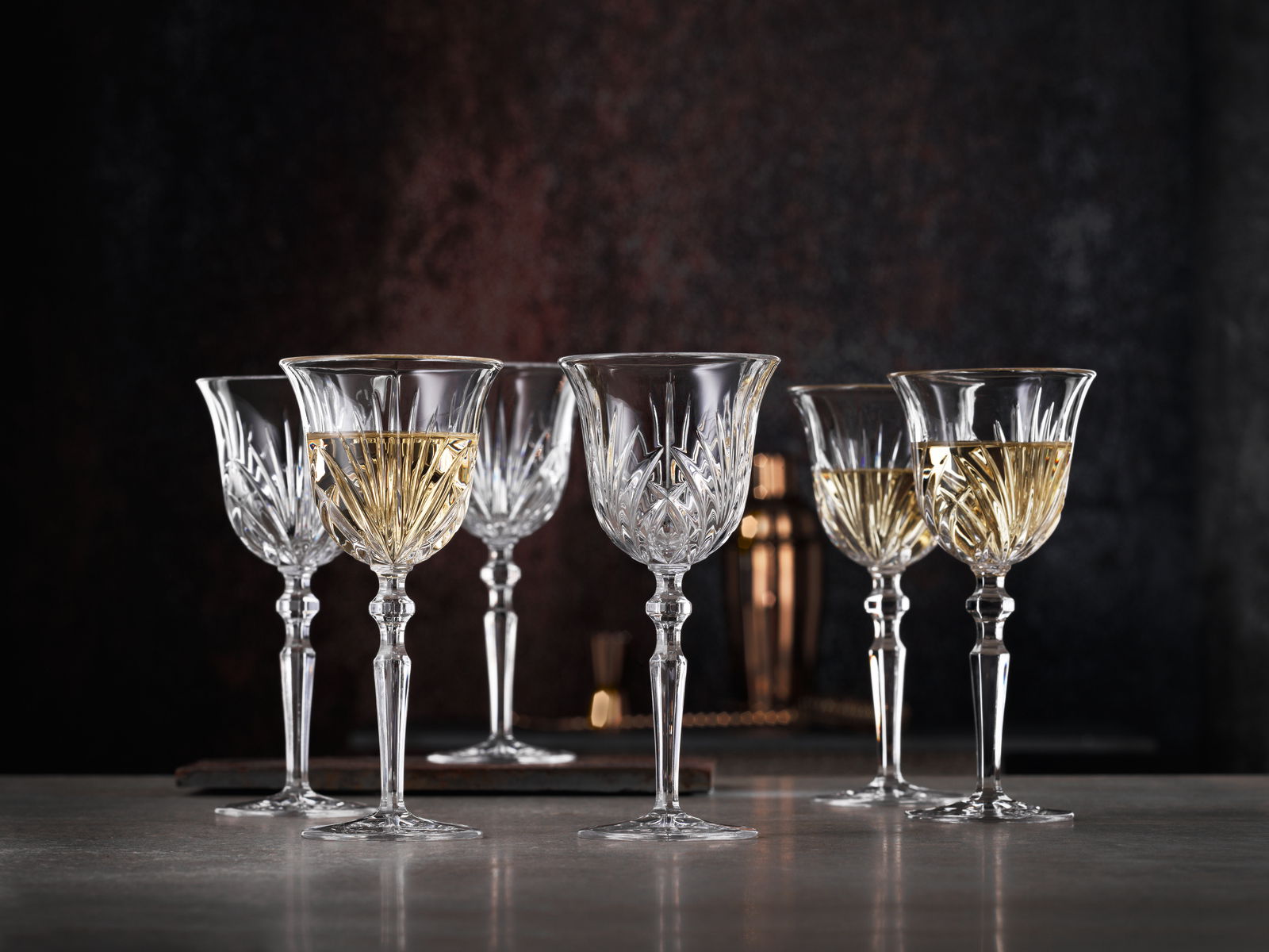 Sechs elegante Weißweinkelche aus Kristallglas mit feinem Schliffmuster, gefüllt mit Weißwein, auf einer Tischoberfläche vor dunklem Hintergrund, seitliche Perspektive.