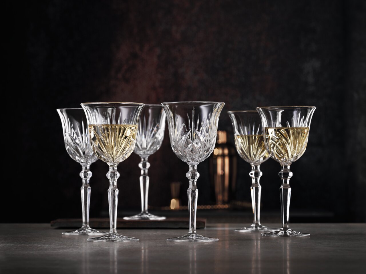 Sechs elegante Weißweinkelche aus Kristallglas mit feinem Schliffmuster, gefüllt mit Weißwein, auf einer Tischoberfläche vor dunklem Hintergrund, seitliche Perspektive.