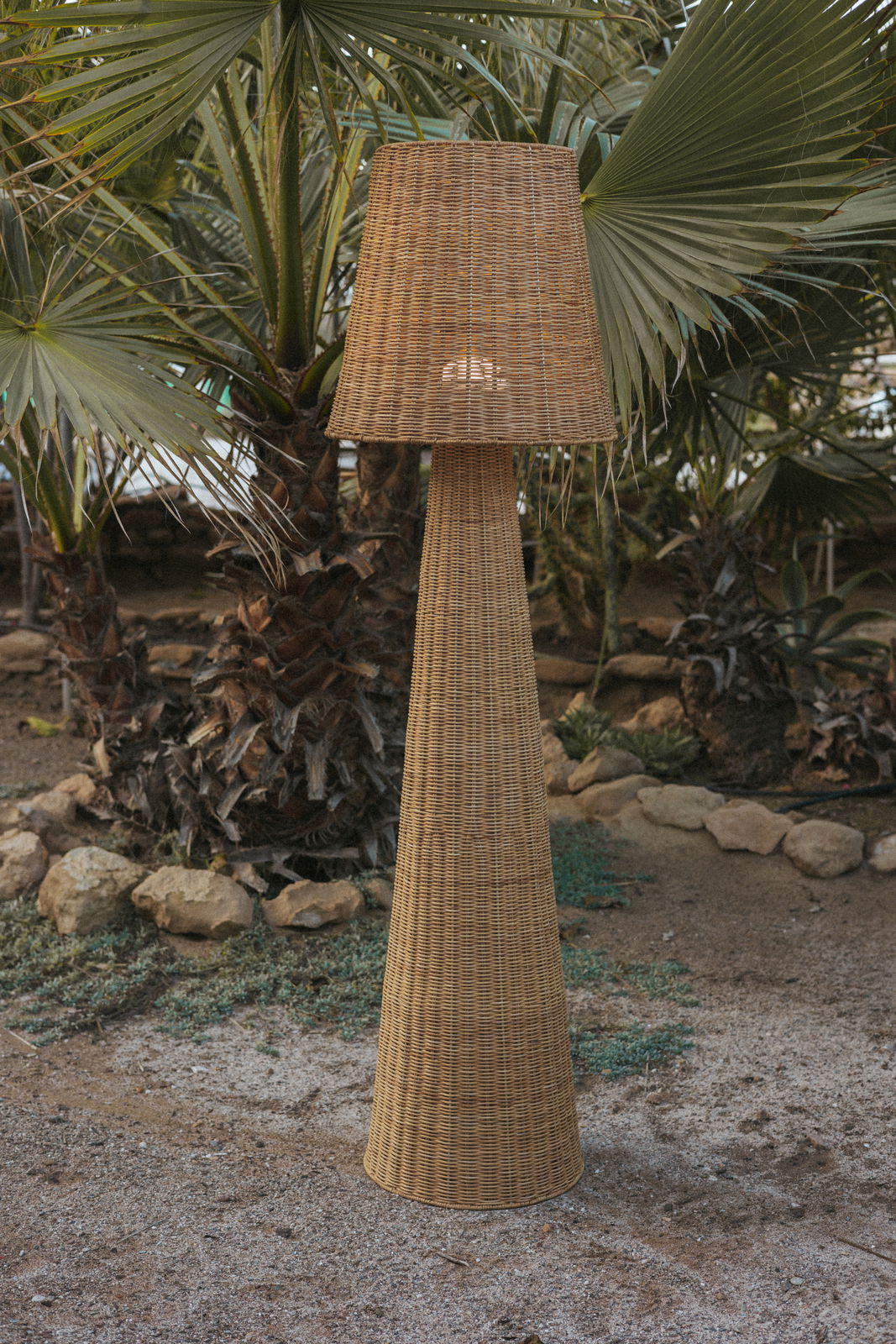 Akku-Outdoor-Stehleuchte aus Rattan in einem Garten mit Palmen, seitliche Perspektive