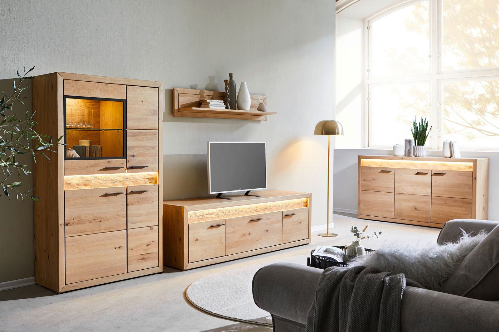 Wohnzimmer mit modernen Holzmöbeln, darunter ein Highboard, ein Lowboard und ein Sideboard, alle mit Beleuchtung. Perspektive von vorne links.