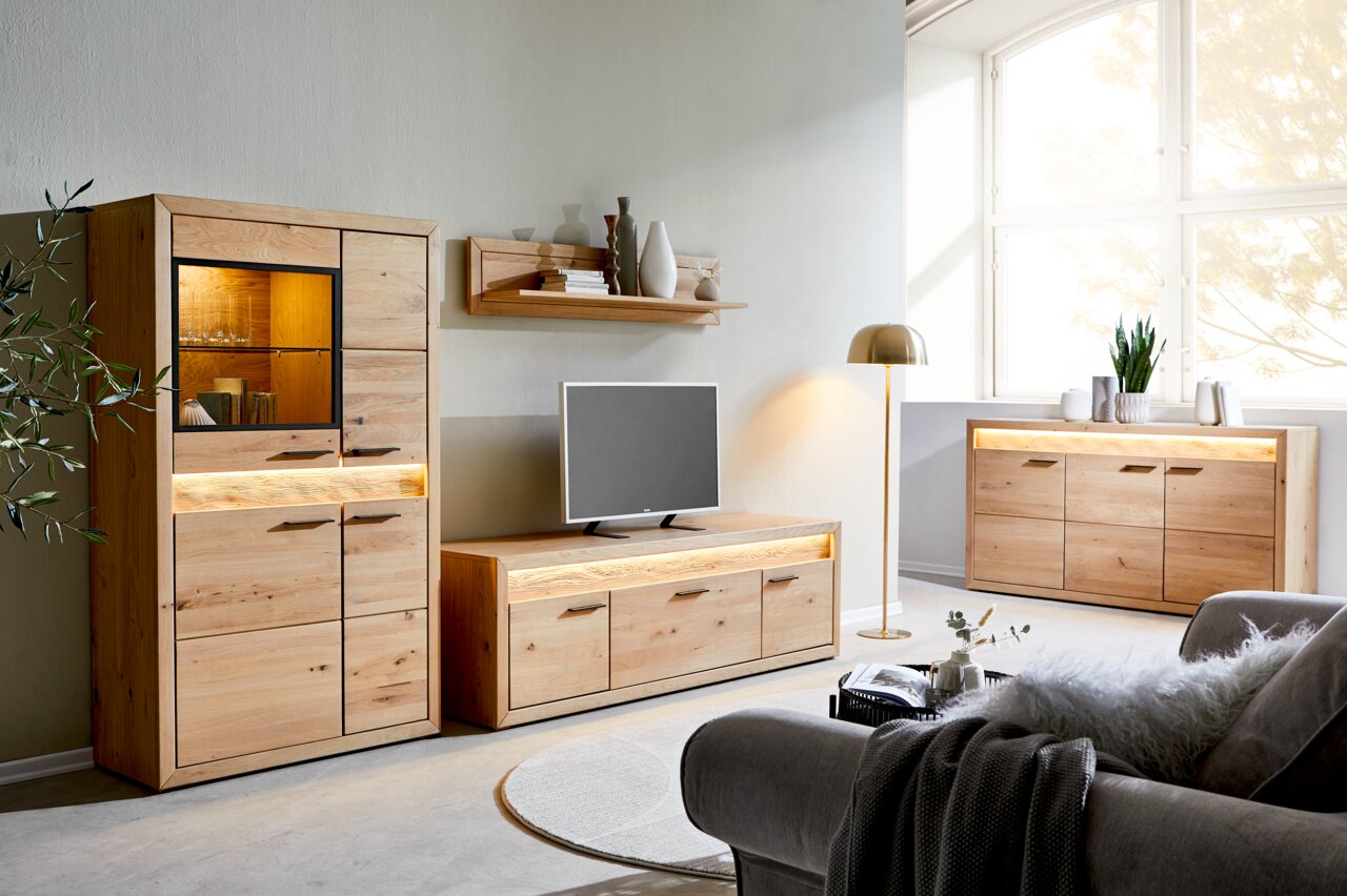 Wohnzimmer mit modernen Holzmöbeln, darunter ein Highboard, ein Lowboard und ein Sideboard, alle mit Beleuchtung. Perspektive von vorne links.