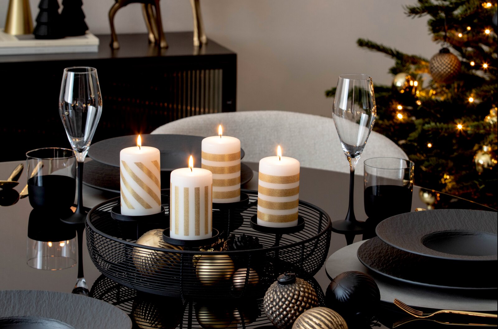 Villeroy & Boch Adventskranz Vier brennende weiße Stumpenkerzen mit goldenen Streifen aus leicht erhöhter Frontalperspektive in einer schwarzen Metallschale auf einer festlich gedeckten Weihnachtstafel