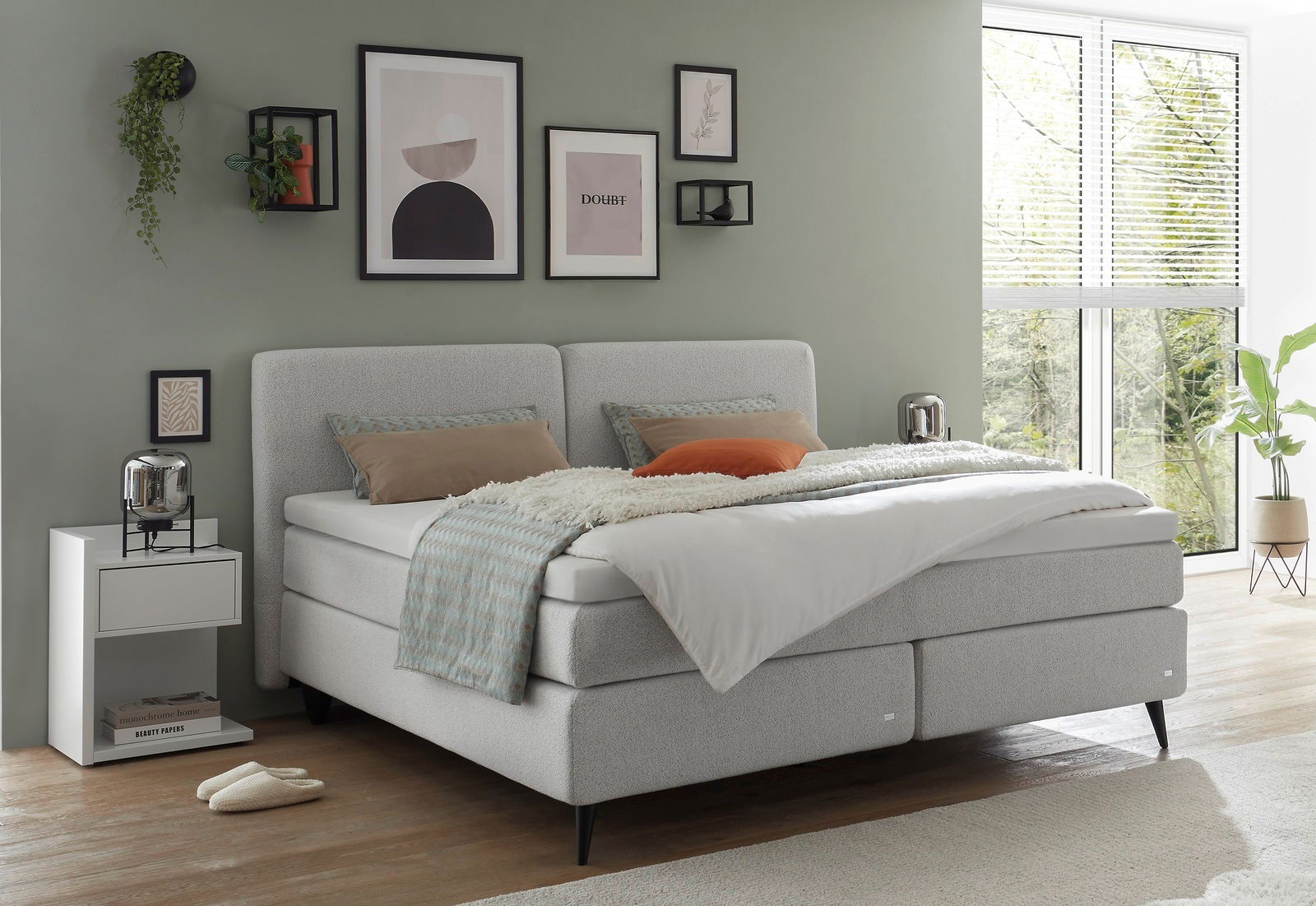 Boxspringbett in einem modernen Schlafzimmer mit grüner Wand, dekorativen Bildern und einem Fenster mit Blick ins Grüne, seitliche Perspektive.