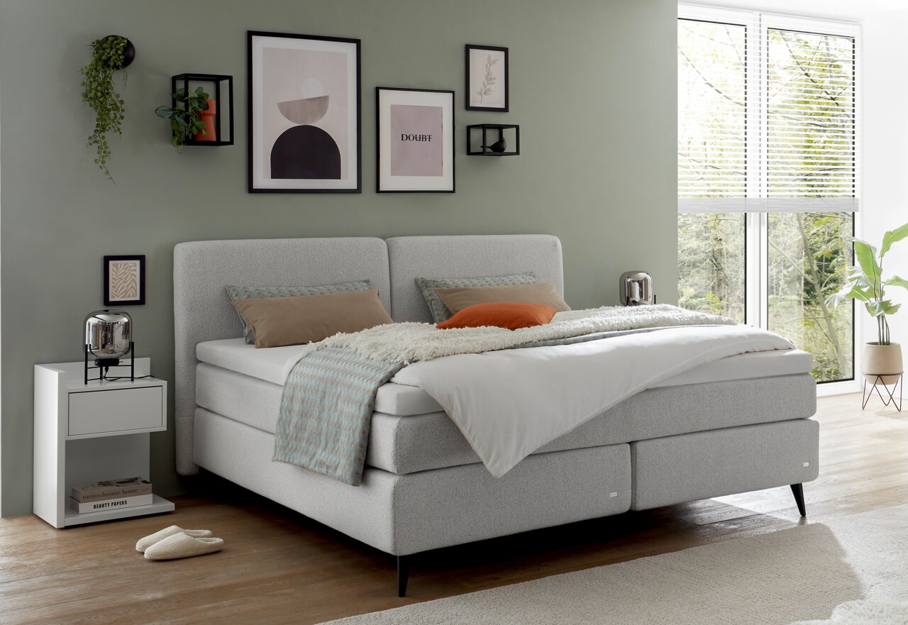 Boxspringbett in einem modernen Schlafzimmer mit grüner Wand, dekorativen Bildern und einem Fenster mit Blick ins Grüne, seitliche Perspektive.