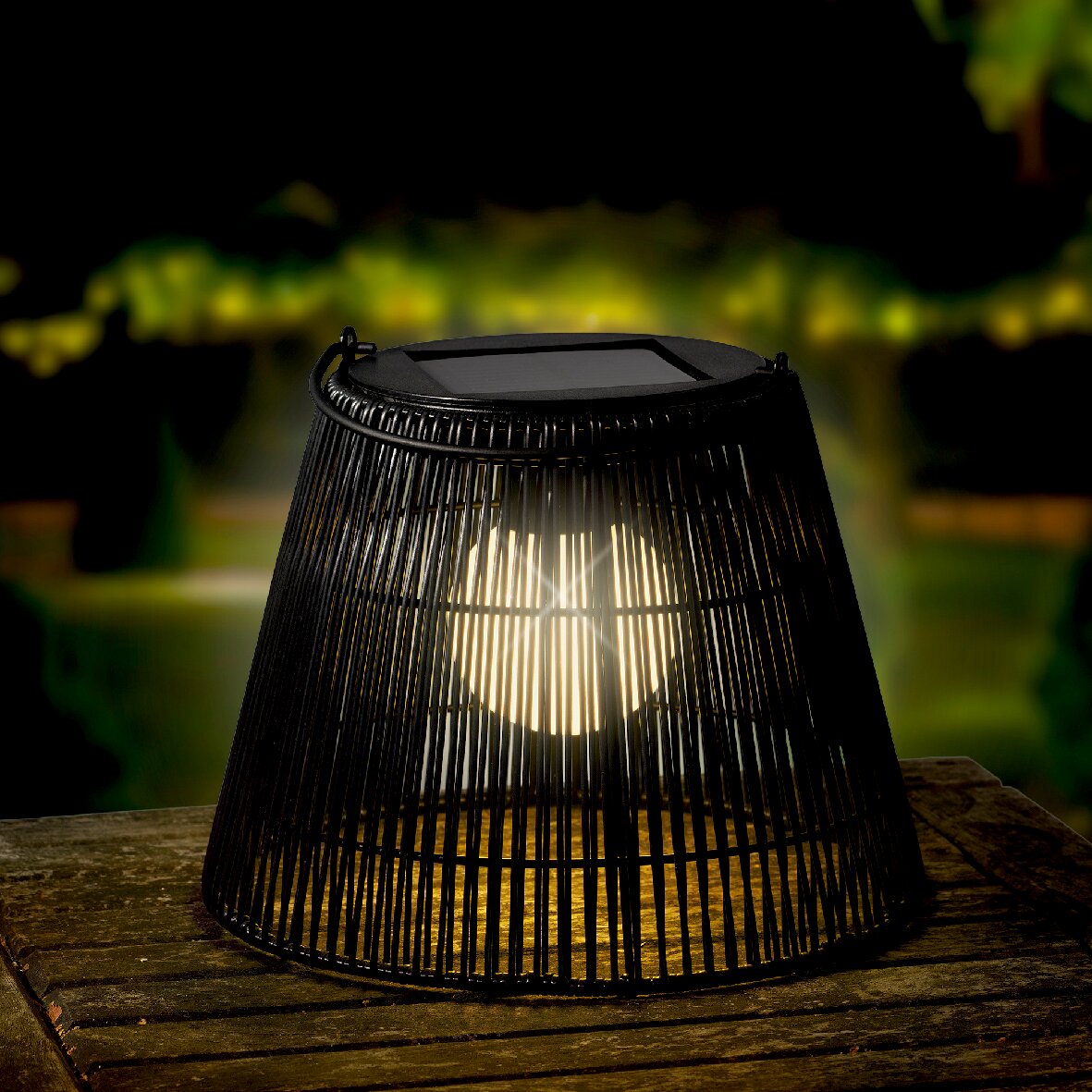 Lumineo Solar Laterne Solarlaterne aus schwarzem Geflecht auf einem Holztisch, leuchtend in der Abenddämmerung, von schräg oben betrachtet.