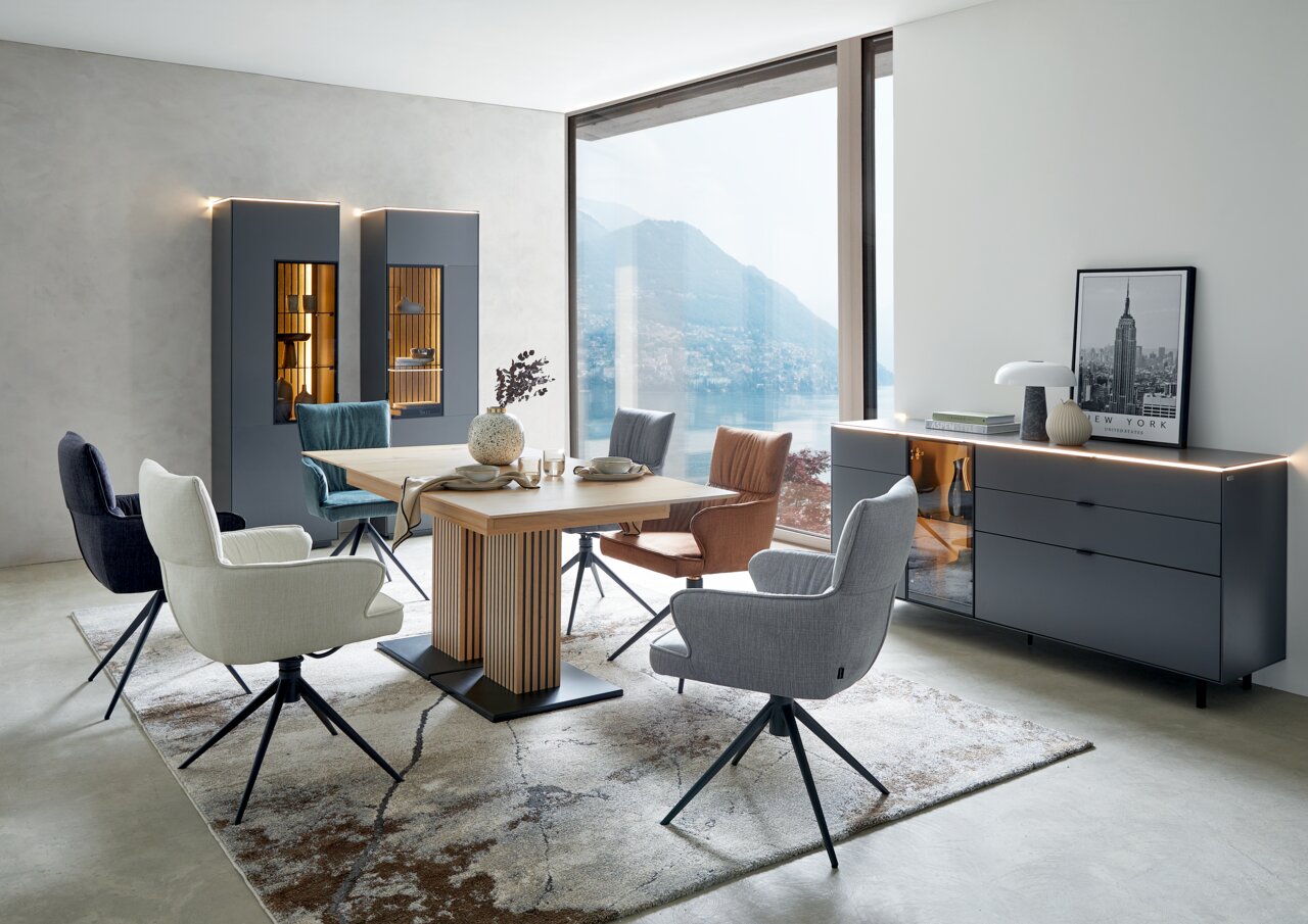 Dieter Knoll Eettafel AVELINO Moderner Essbereich mit einem großen Holztisch und verschiedenen gepolsterten Stühlen in einem hellen Raum mit Blick auf einen See.