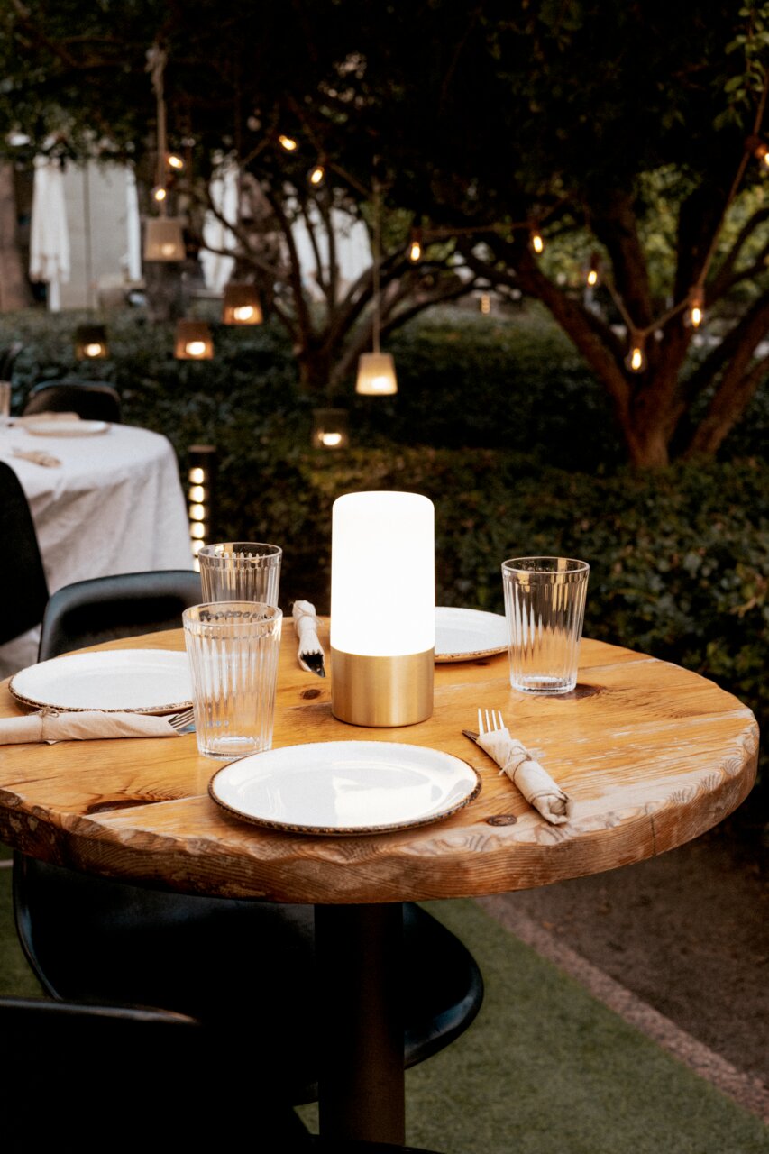 Leuchtende, zylindrische Tischlampe mit mattem Diffusor und goldfarbenem Sockel auf rundem Holztisch im Outdoor-Restaurant, umgeben von Tellern, Gläsern und Besteck; Perspektive: leicht erhöhte Frontansicht.