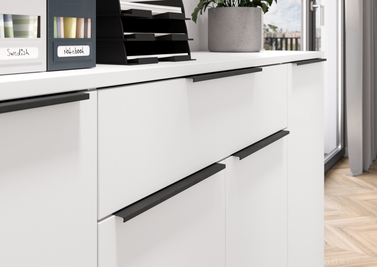 Nahaufnahme eines weißen Sideboards mit schwarzen Griffen, darauf stehen Aktenablagen und eine Pflanze, seitliche Perspektive.