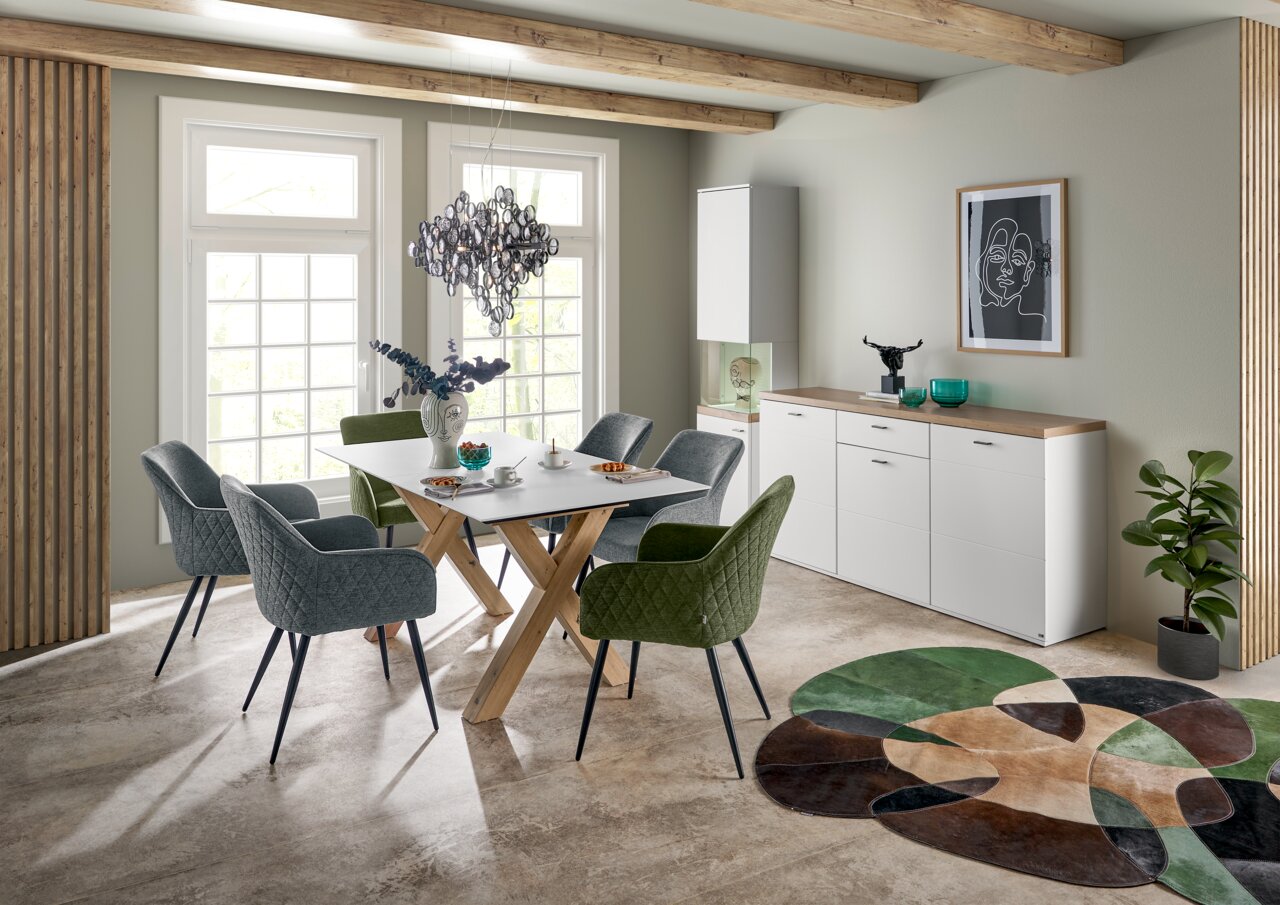 Moderner Esstisch mit Holzkreuzgestell und weißer Tischplatte, umgeben von gepolsterten Stühlen in einem stilvollen Esszimmer mit Blick auf große Fenster.