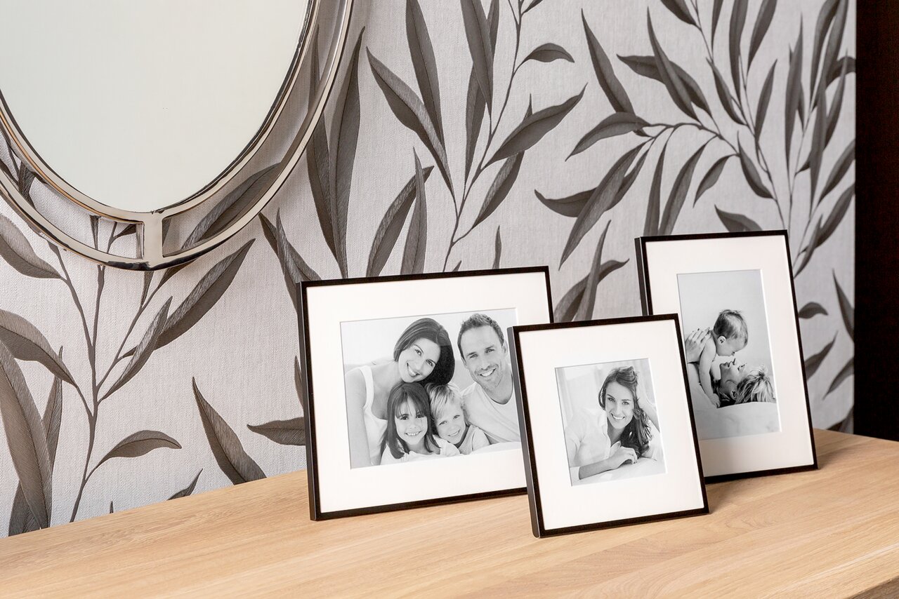 Drei schwarze Aluminiumrahmen auf einem Holztisch, mit Familienfotos, vor einer Wand mit Blattmuster, aus seitlicher Perspektive.