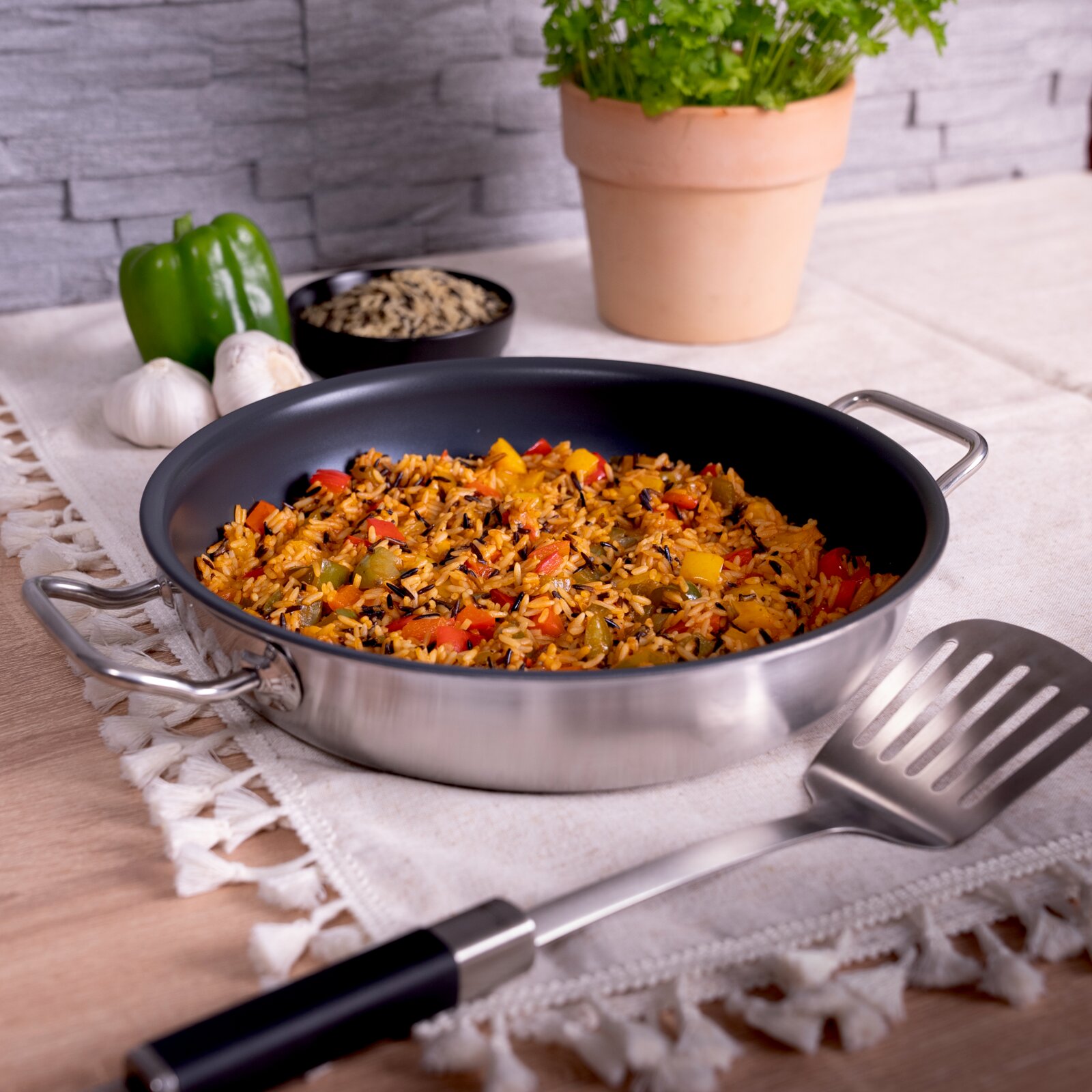 Schuine bovenaanzicht op een roestvrijstalen hapjespan met twee handgrepen, gevuld met gekruide rijst met paprika, op een keukentafel met spatel en verse ingrediënten op de achtergrond.