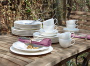 Ein 30-teiliges weißes Geschirrset auf einem Holztisch im Freien, bestehend aus Tellern, Tassen und Untertassen, mit einer lila Serviette und einem Namensschild. Perspektive von schräg oben.