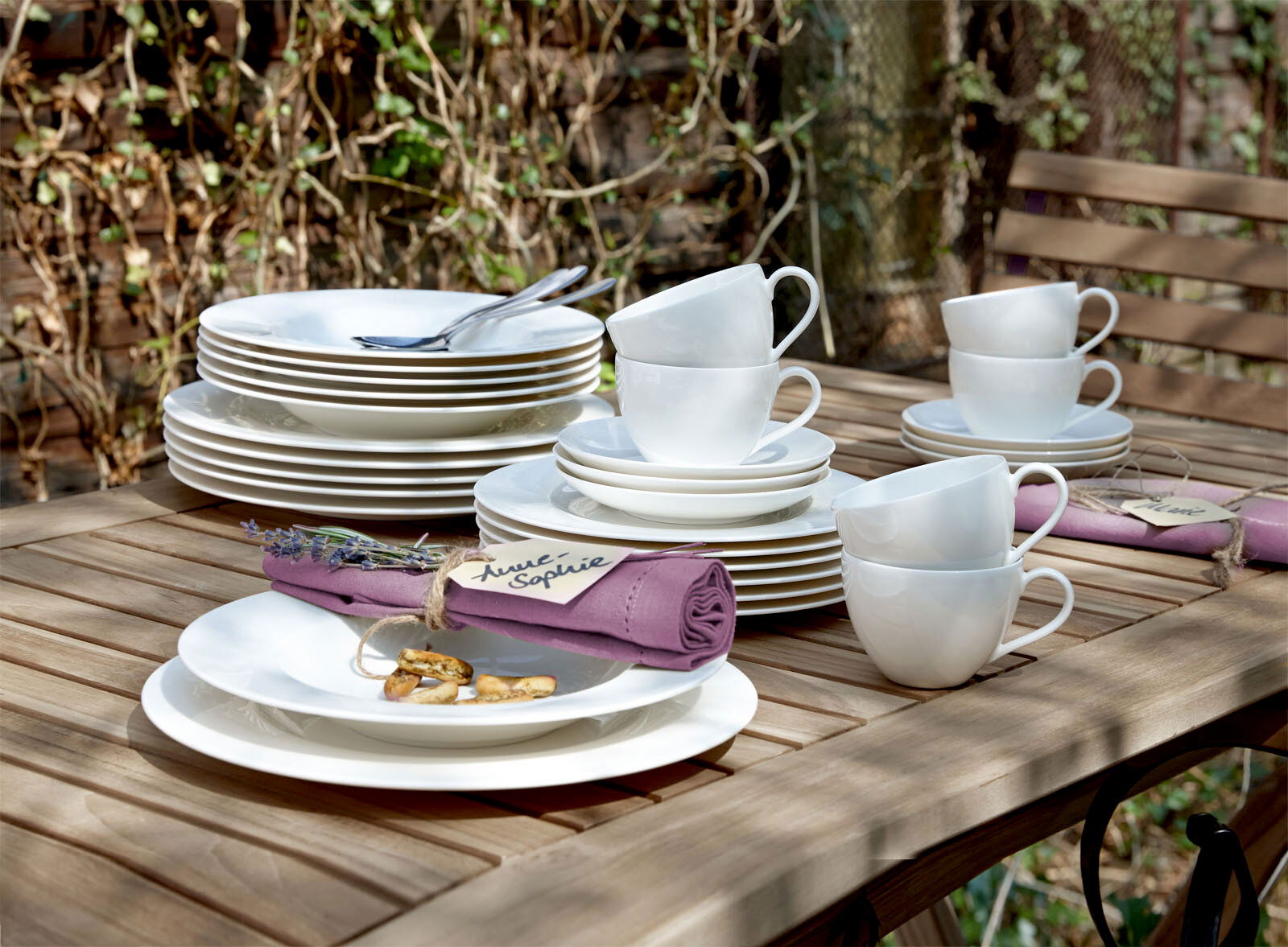 Ein 30-teiliges weißes Geschirrset auf einem Holztisch im Freien, bestehend aus Tellern, Tassen und Untertassen, mit einer lila Serviette und einem Namensschild. Perspektive von schräg oben.