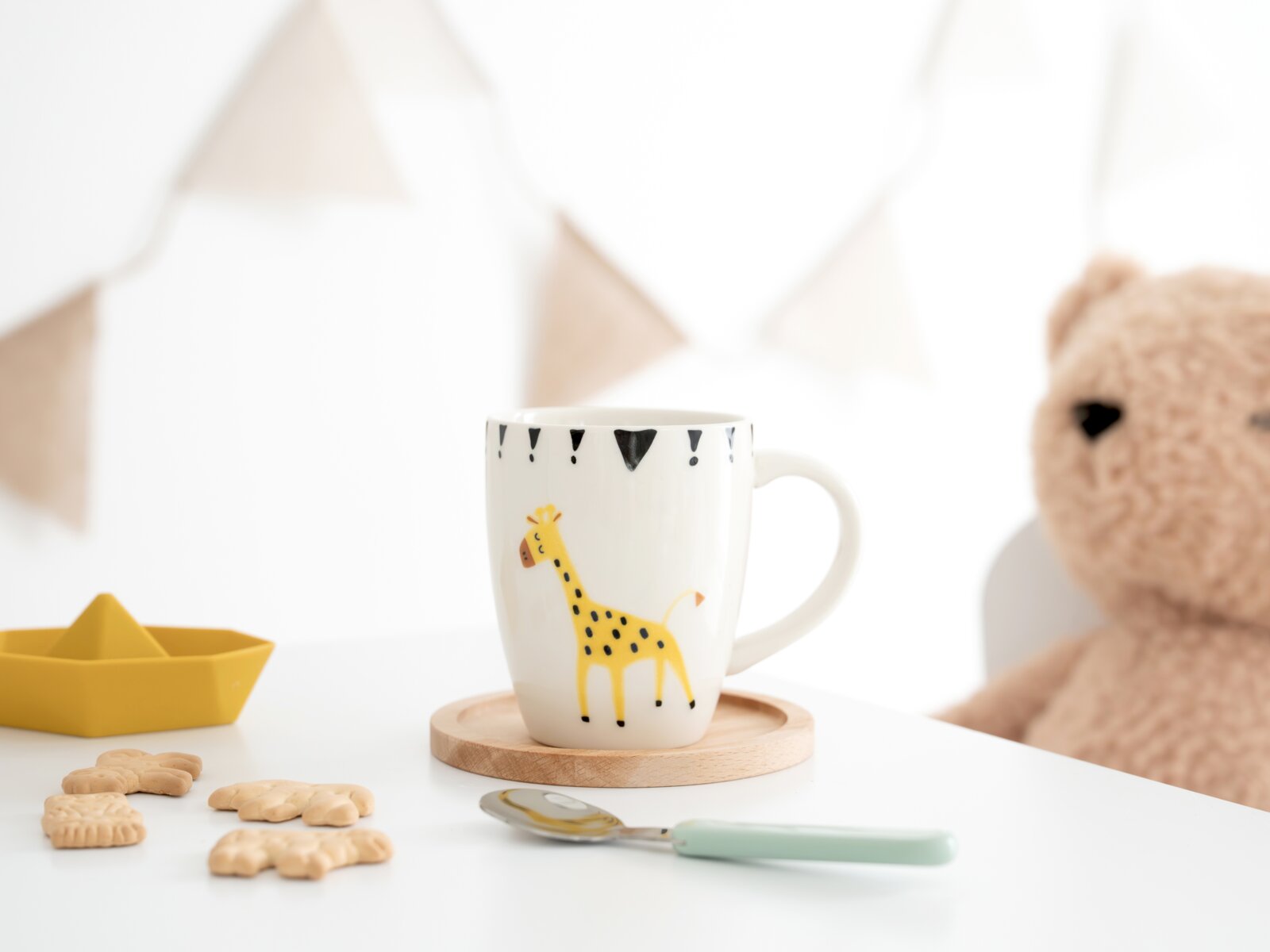 Weiße Kindertasse mit gelbem Giraffenmotiv und schwarzen Dreiecken, frontal leicht von oben auf einem Holzuntersetzer fotografiert, umgeben von Keksen, Löffel, Spielzeugboot und Teddybär.
