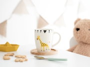 Weiße Kindertasse mit gelbem Giraffenmotiv und schwarzen Dreiecken, frontal leicht von oben auf einem Holzuntersetzer fotografiert, umgeben von Keksen, Löffel, Spielzeugboot und Teddybär.