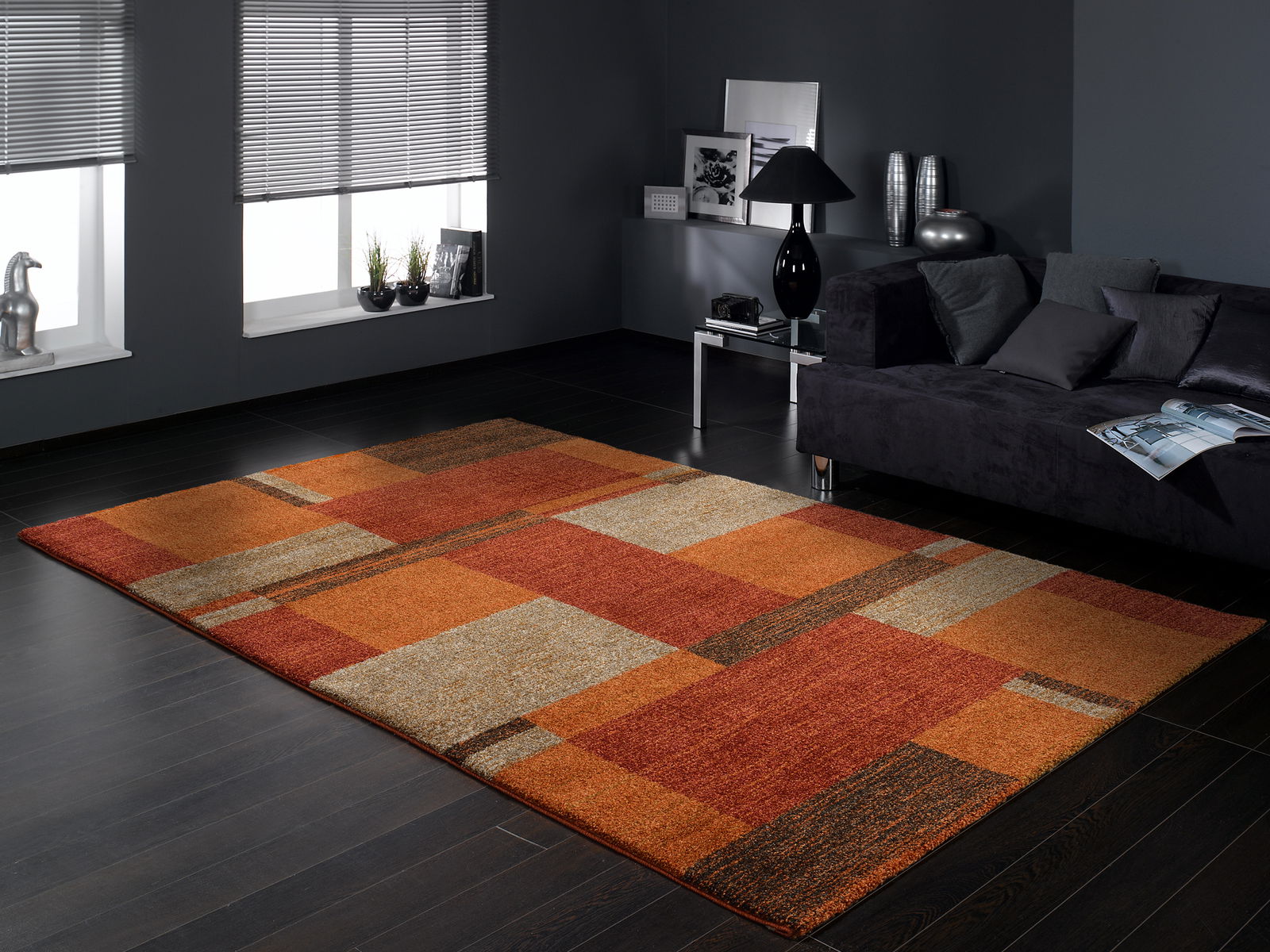 Frisé-Teppich in warmen Rot- und Orangetönen mit geometrischem Muster, in einem modernen Wohnzimmer mit dunklem Holzboden und grauem Sofa, aus der Perspektive von schräg oben fotografiert.