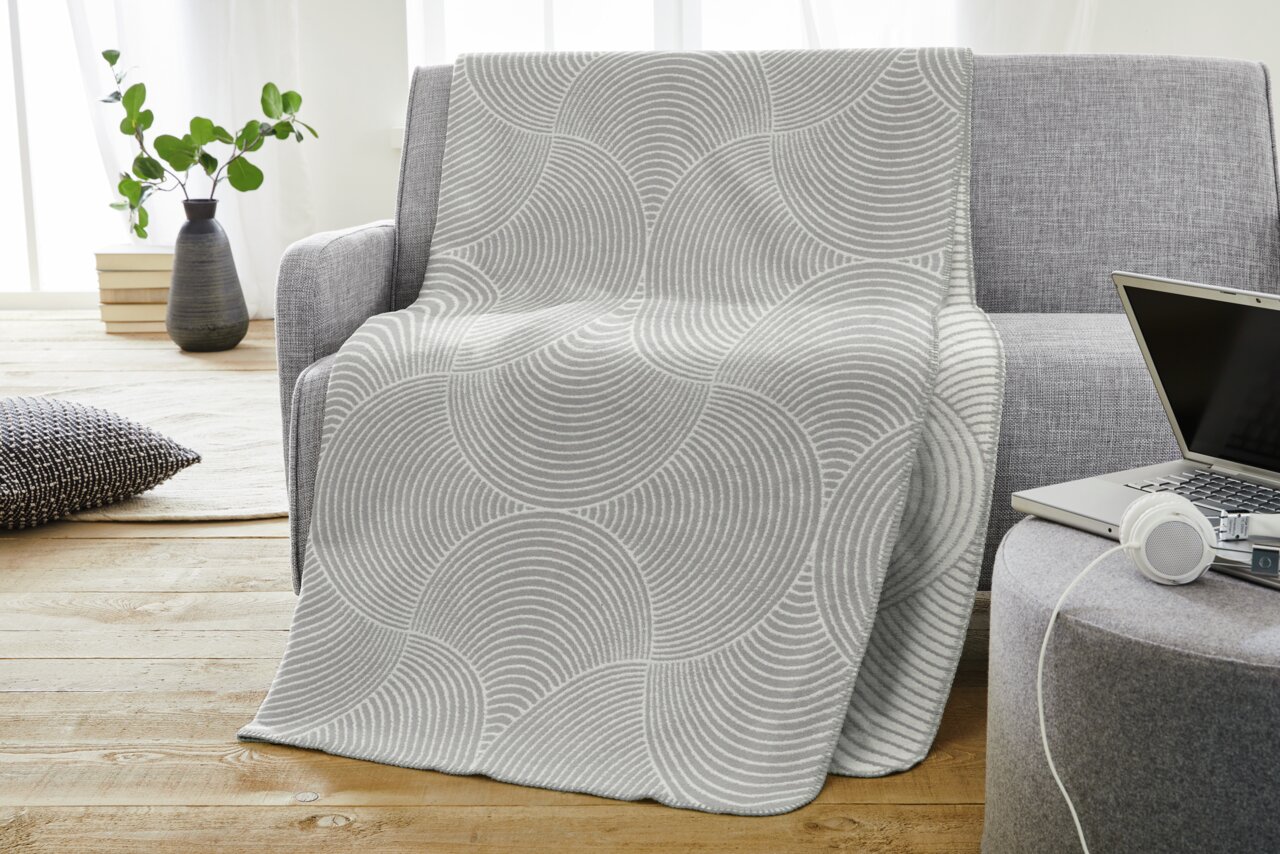 Graue Jacquard Decke mit kreisförmigem Muster auf einem Sofa drapiert, seitliche Perspektive.