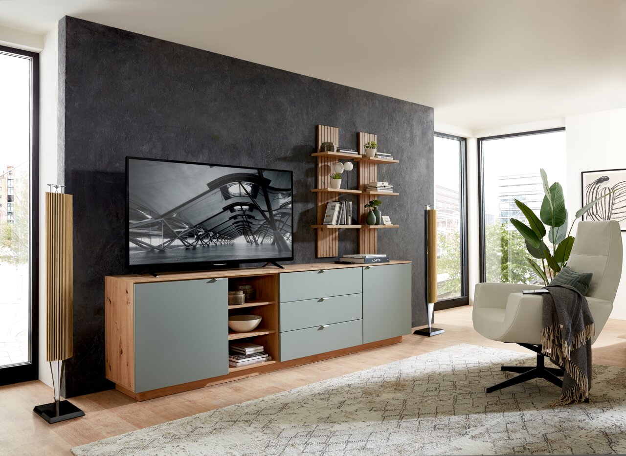 Modernes Wohnzimmer mit einem Wandpaneel im Hintergrund, das mit Regalen dekoriert ist. Perspektive von der Seite, die den Raum mit einem Fernseher, einem Sideboard und einem Sessel zeigt.