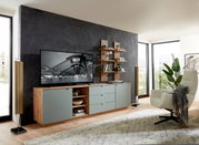 Modernes Wohnzimmer mit einem Wandpaneel im Hintergrund, das mit Regalen dekoriert ist. Perspektive von der Seite, die den Raum mit einem Fernseher, einem Sideboard und einem Sessel zeigt.
