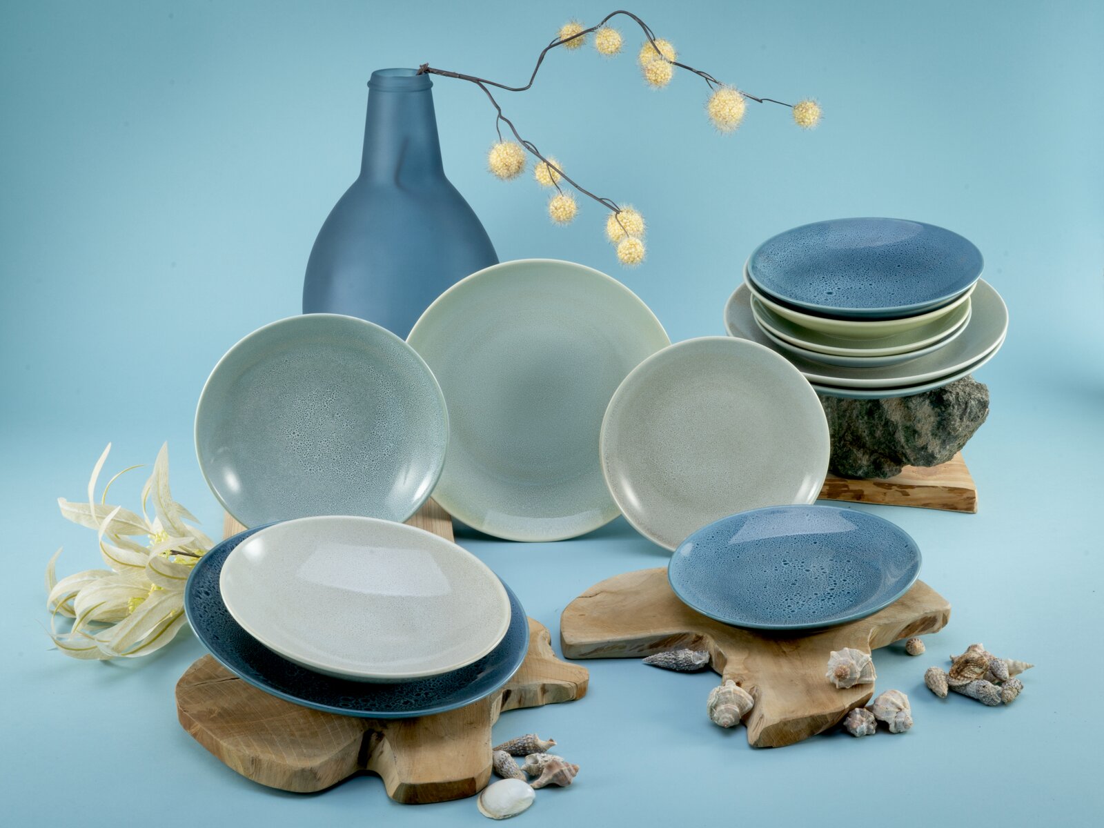 Keramik-Geschirrset in Blau- und Grautönen mit Tellern und Schalen, dekorativ arrangiert mit blauer Vase, Lichterkette, Holzuntersetzern und Muscheln vor hellblauem Hintergrund, fotografiert in frontaler Studio-Perspektive mit leicht erhöhter Ansicht
