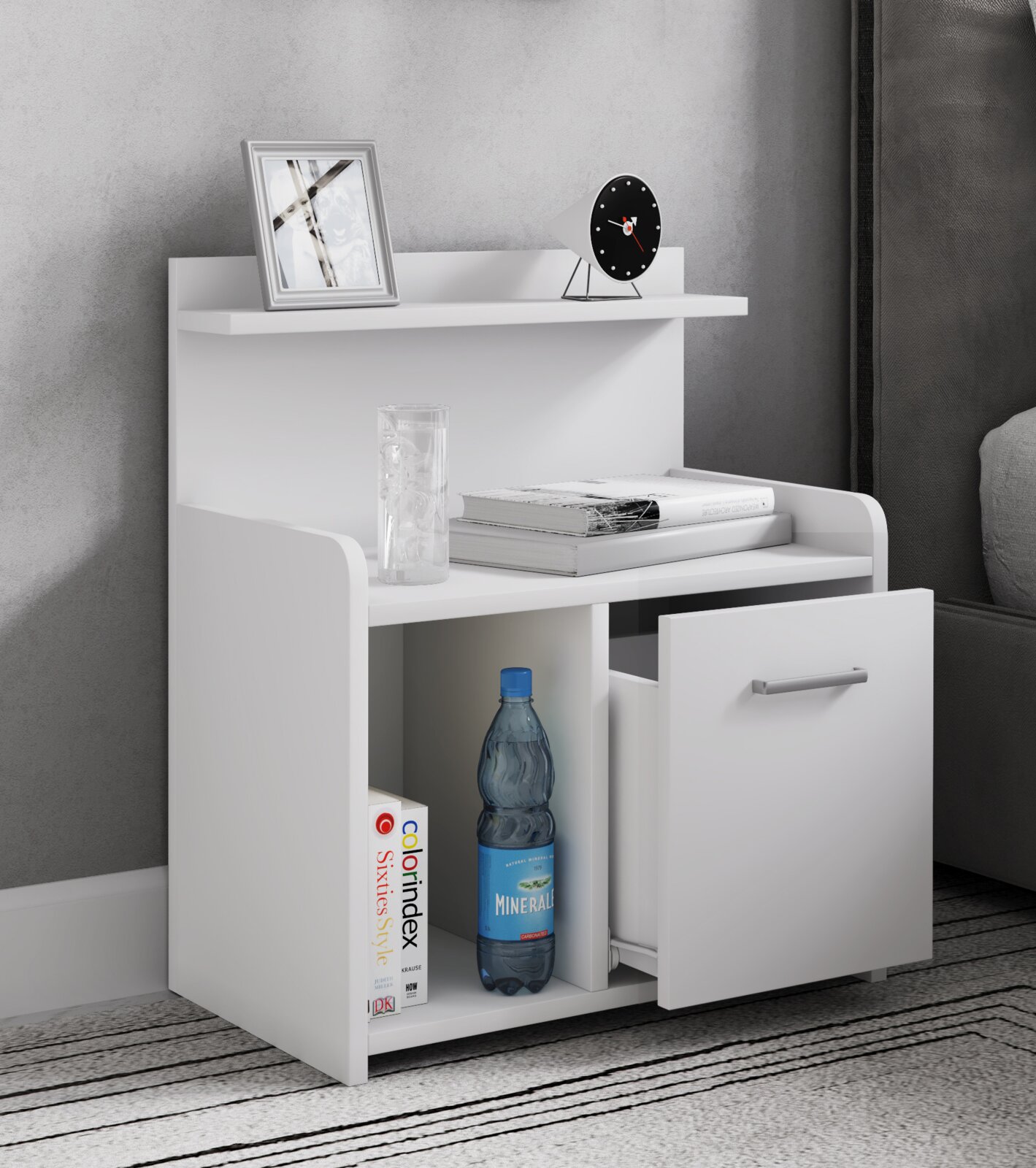 Weißer moderner Nachttisch mit Ablage, offenem Fach und geöffneter Schublade mit Metallgriff, dekoriert mit Bilderrahmen, kleiner Uhr, Büchern und Wasserflasche, fotografiert in leichter Frontansicht von schräg rechts.
