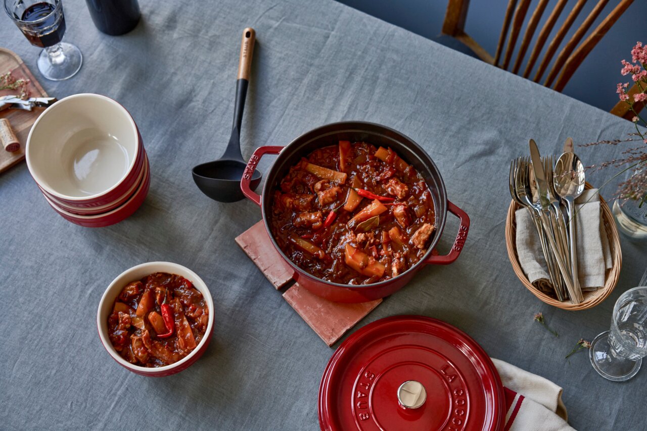 Rote Cocotte 24cm rund von oben mit Eintopf gefüllt, auf einem gedeckten Tisch mit Schalen und Besteck.