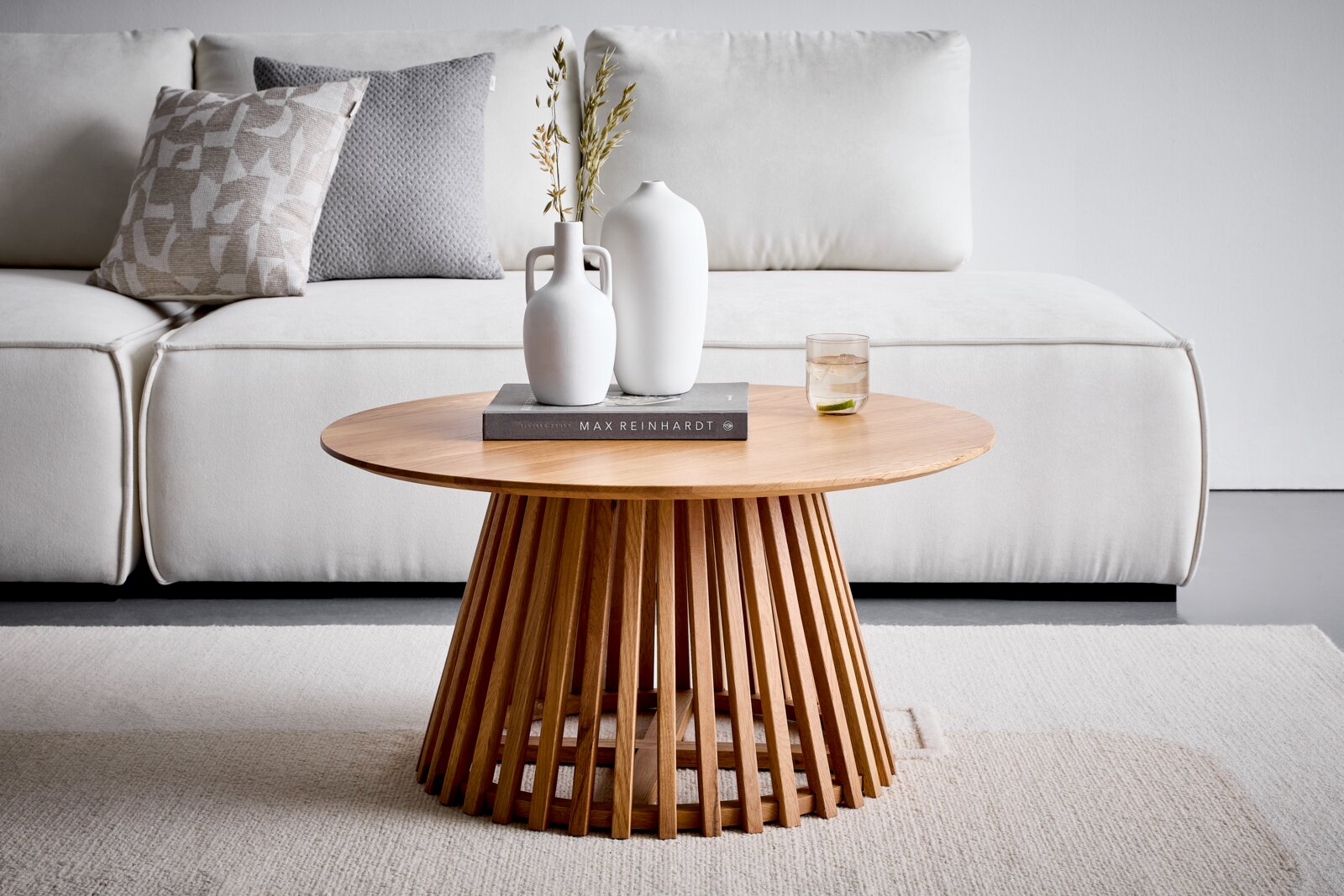 Cantus Couchtisch NARVIK Runder Couchtisch aus hellem Holz mit geriffeltem Lamellenfuß, frontal auf Augenhöhe im Wohnzimmer vor weißem Sofa, dekoriert mit zwei weißen Vasen, Buch und Glas