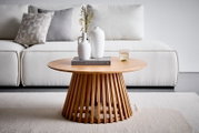 Runder Couchtisch aus hellem Holz mit geriffeltem Lamellenfuß, frontal auf Augenhöhe im Wohnzimmer vor weißem Sofa, dekoriert mit zwei weißen Vasen, Buch und Glas