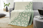 Jacquard Decke s.Oliver in hellem Beige mit grünem Buchstabenmuster, drapiert über einem grauen Sofa in einem modernen Wohnzimmer.