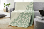 Jacquard Decke s.Oliver in hellem Beige mit grünem Buchstabenmuster, drapiert über einem grauen Sofa in einem modernen Wohnzimmer.