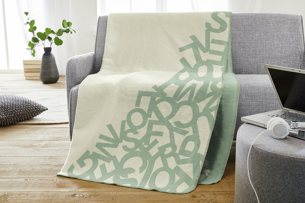 s.Oliver Wohndecke 150x200cm weiß/grün Jacquard Decke s.Oliver in hellem Beige mit grünem Buchstabenmuster, drapiert über einem grauen Sofa in einem modernen Wohnzimmer.