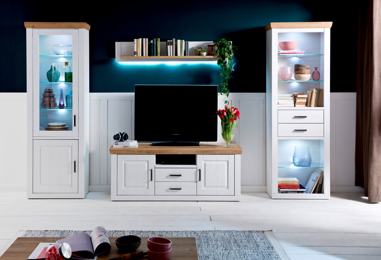 Wandmeubelset in landhuisstijl in wit met accenten in houtlook, bestaande uit een tv-lowboard, twee vitrinekasten met ledverlichting en een wandplank, gefotografeerd vanaf de voorkant in een woonkamer