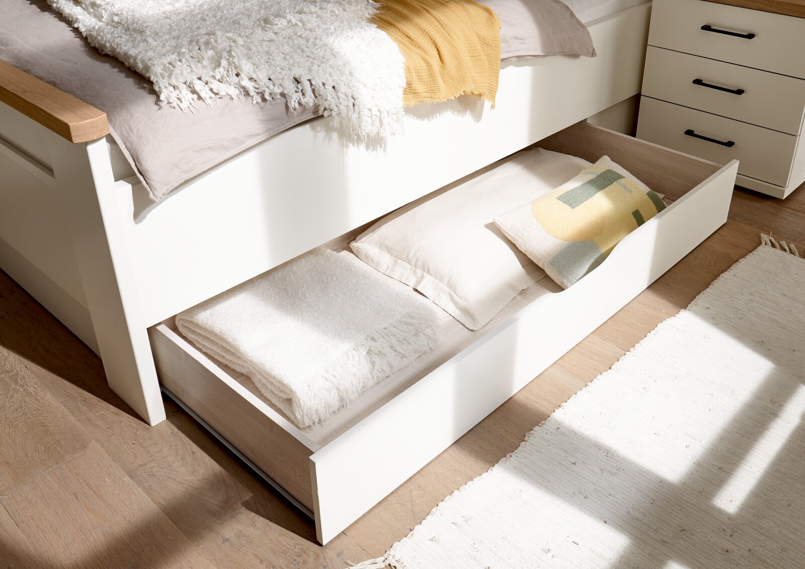 Weißes Bett mit ausgezogenem Unterbett-Schubkasten zur Aufbewahrung von Decken und Kissen, seitliche Perspektive in hellem Schlafzimmer