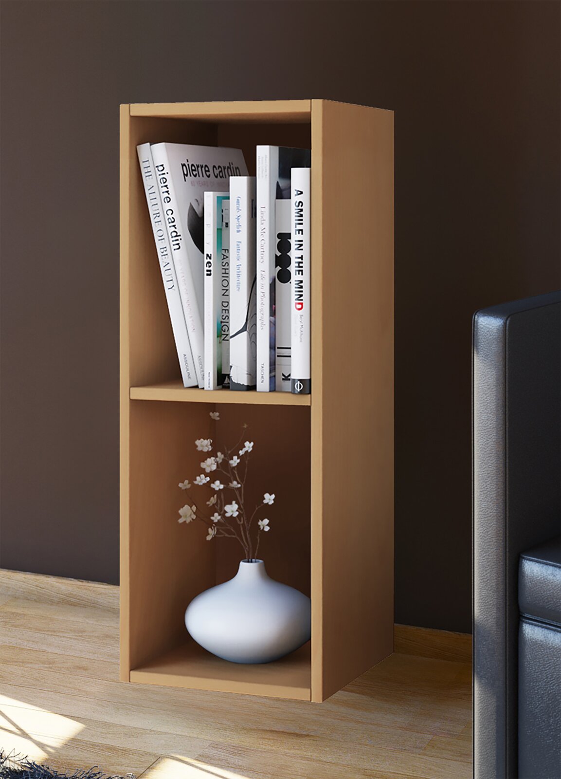 Schmales, offenes Bücherregal aus hellem Holz mit zwei Fächern, oben mit Büchern und unten mit weißer Vase mit Zweigen, im Wohnzimmer neben einem Sofa, Perspektive: frontale Ansicht leicht von rechts.