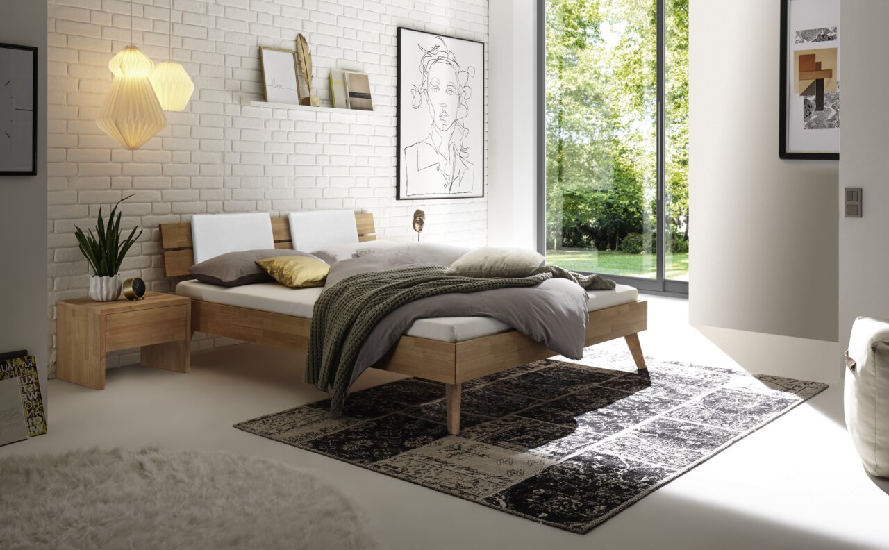 Modernes Schlafzimmer mit einem Kopfteil aus Holz und Metall, seitliche Perspektive. Das Bett ist mit neutralen Farben dekoriert und steht vor einer weißen Ziegelwand mit Kunstwerken und Pflanzen.