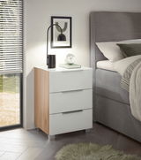 Moderne Nachtkommode mit drei Schubladen in Weiß und Holzoptik, seitliche Perspektive neben einem Bett, dekoriert mit einer Lampe und einem Buch.