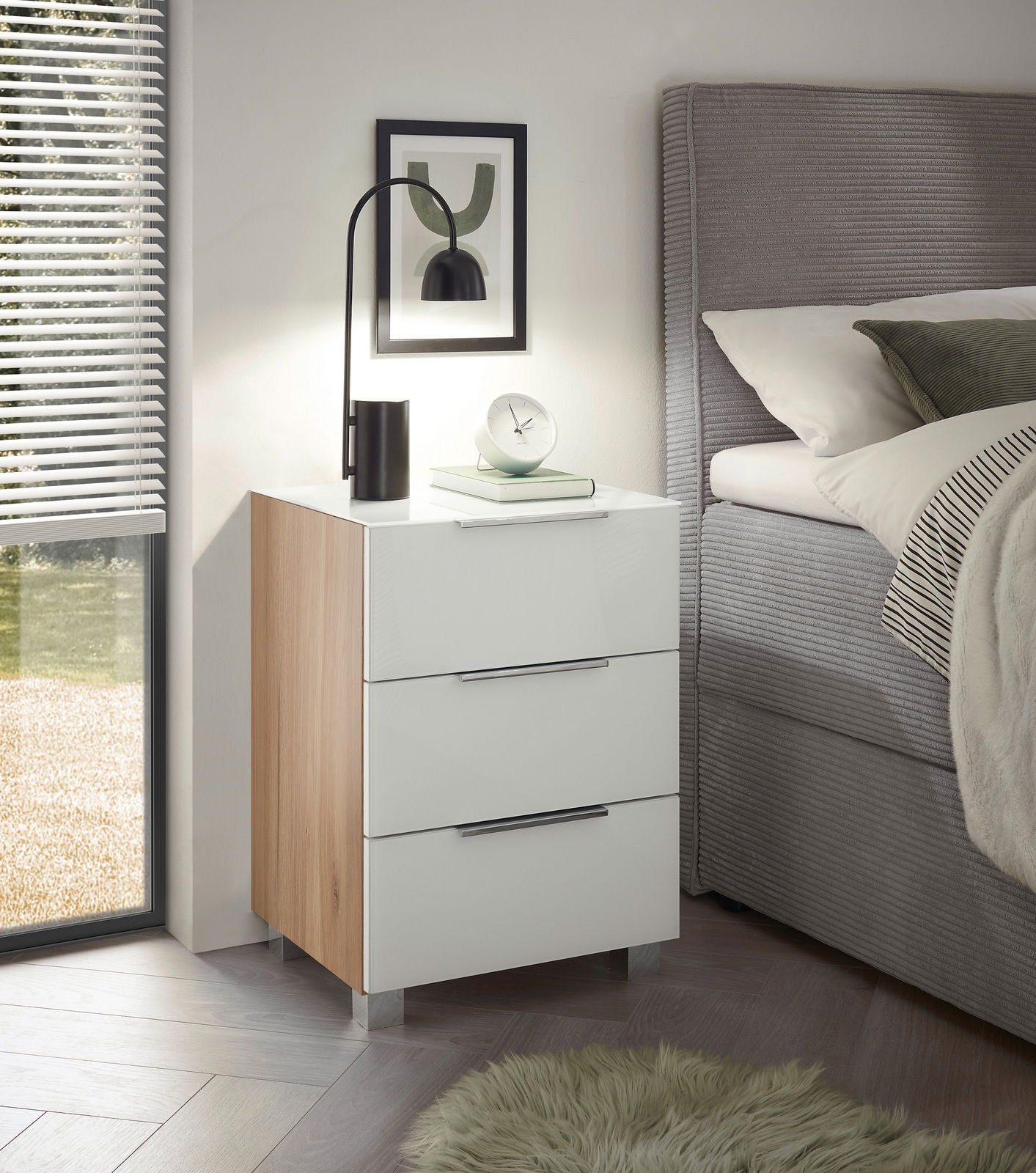 Moderne Nachtkommode mit drei Schubladen in Weiß und Holzoptik, seitliche Perspektive neben einem Bett, dekoriert mit einer Lampe und einem Buch.