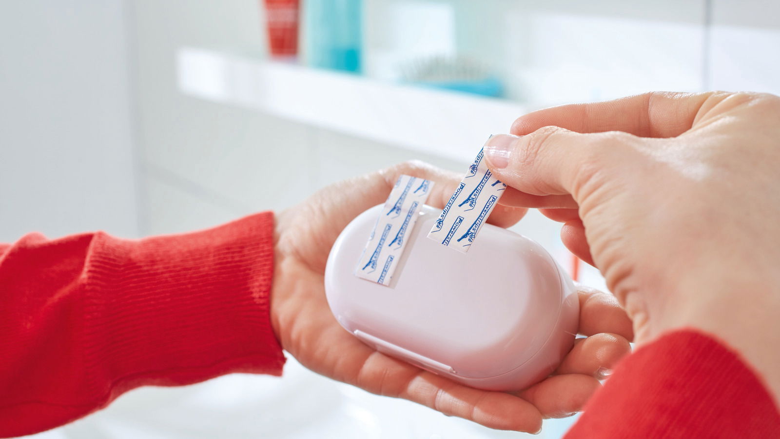 Nahaufnahme von Händen, die tesa Powerstrips Waterproof auf einer weißen Seifenschale anbringen, im Hintergrund ein Badezimmerregal.