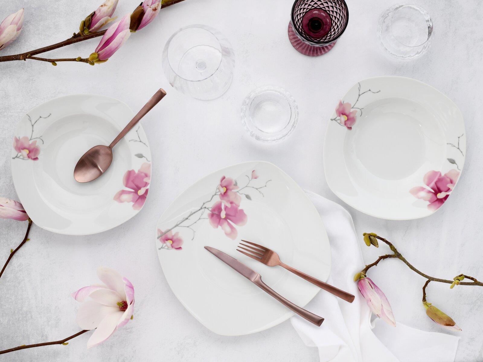 Draufsicht auf weißes Porzellan-Geschirr mit rosa Blütenmotiv: drei Teller/Schalen, roségoldenes Besteck, Gläser und Magnolienzweige auf hellem Tischgedeck.