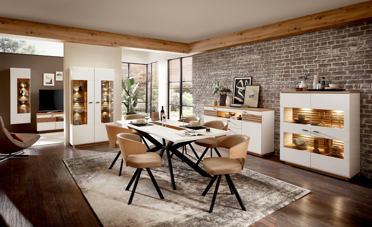 Zurbrüggen Dressoir FRESNO Moderne woonkamermeubels in een stijlvolle ruimte met eettafel, stoelen en verlichte hoge kasten, gezien vanuit de zijkant.