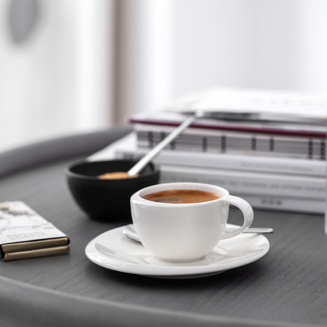 Weiße Espressotasse NewMoon 50ml auf Untertasse, gefüllt mit Espresso, auf einem Tisch mit unscharfem Hintergrund von Büchern und einer schwarzen Schale. Seitliche Perspektive.