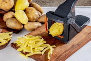 Zwarte frietsnijder met hendel snijdt een geschilde aardappel in frieten op een houten snijplank, met aardappelen op de achtergrond; close-up vanuit schuin bovenaanzicht.