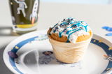 Kindergeschirr mit Weltall-Motiv, bestehend aus einem Teller mit Astronauten- und Raketenillustrationen, im Vordergrund ein dekorierter Muffin in einer Waffel, seitliche Perspektive.