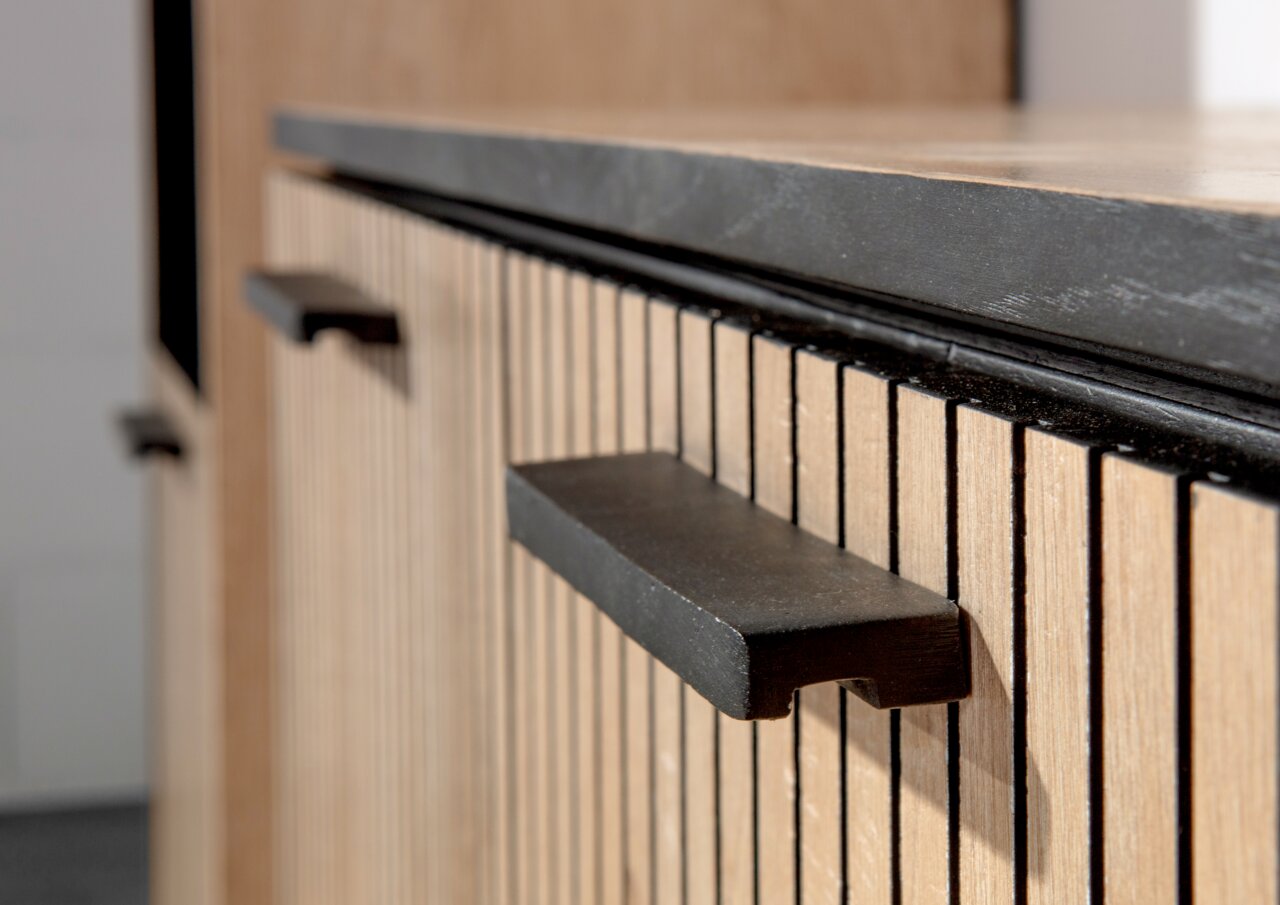 Nahaufnahme eines Möbelstücks mit vertikalen Holzlamellen und schwarzen Griffen, seitliche Perspektive