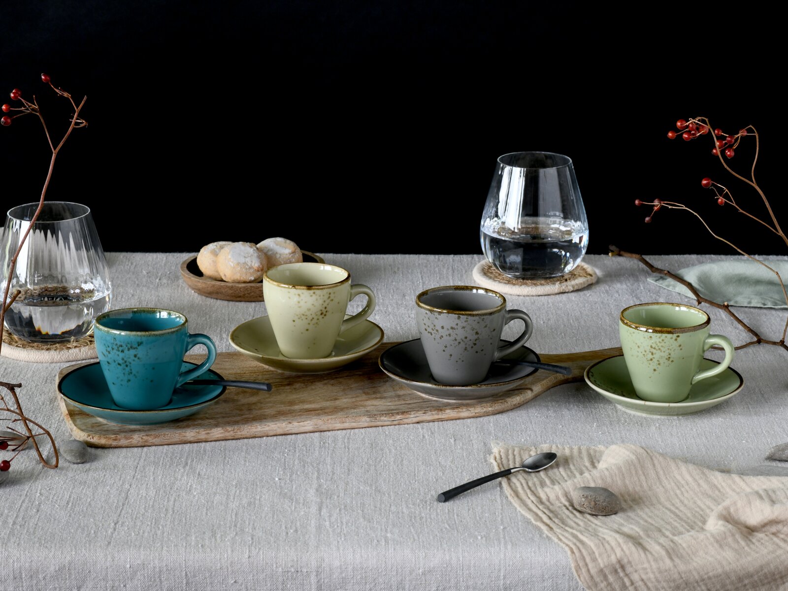 Schräg von vorn fotografiertes Kaffee-Set: vier gesprenkelte Keramik-Tassen mit Untertassen in Blau, Creme, Grau und Grün auf Holzbrett, dazu Wassergläser und Gebäck auf grauer Tischdecke vor schwarzem Hintergrund.