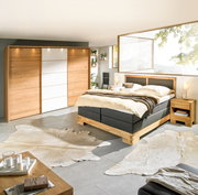 Modernes Schlafzimmer mit Boxspringbett, Nachtkommode und großem Kleiderschrank aus Holz, seitliche Perspektive.