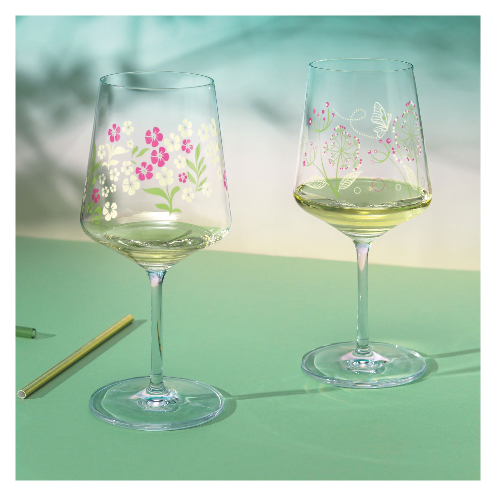Zwei Weingläser mit floralen Mustern, eines mit rosa und weißen Blumen, das andere mit Schmetterling und Blättern, auf einer grünen Oberfläche aus seitlicher Perspektive.