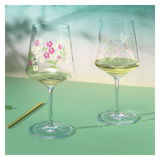 Zwei Weingläser mit floralen Mustern, eines mit rosa und weißen Blumen, das andere mit Schmetterling und Blättern, auf einer grünen Oberfläche aus seitlicher Perspektive.