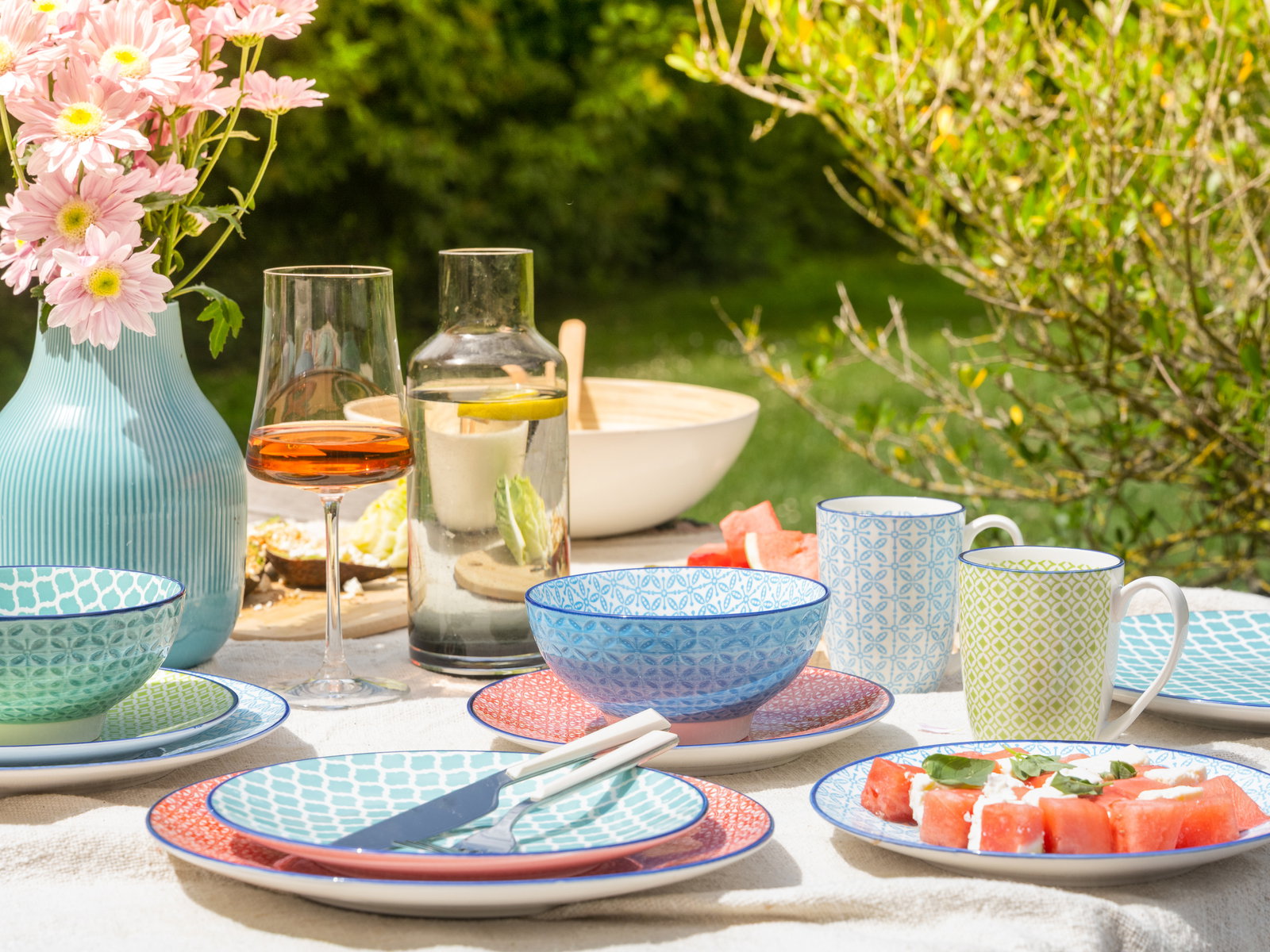 Ein farbenfrohes Kombiservice-Set mit Tellern, Schalen und Tassen in verschiedenen Mustern, arrangiert auf einem Tisch im Freien mit Blumen und Getränken, aus der Vogelperspektive fotografiert.